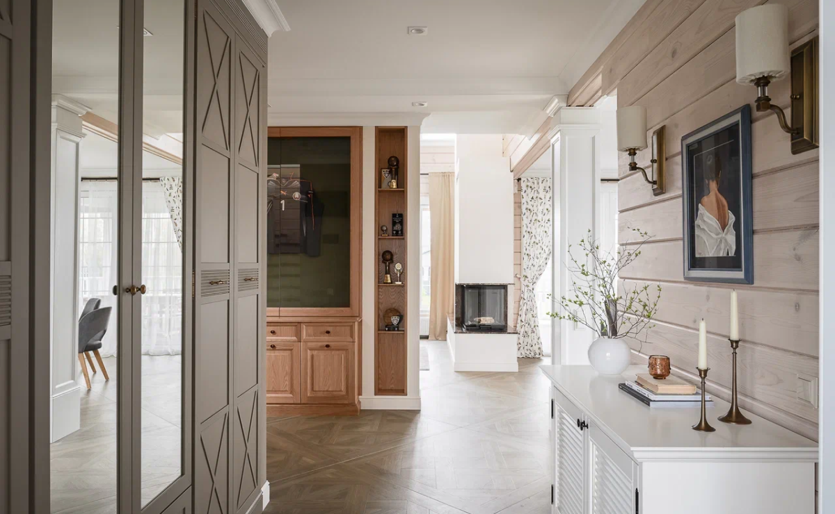 На первом этаже мы разместили прихожую и коридор, хозяйственную зону, кухню-столовую, гостевой санузел, гостиную и игровую.<br>
 На втором – расположены хозяйская спальня с гардеробной и санузлом, гостевая комната, детская спальня. В большом холле придумана зона для уединения и релакса с мягкой мебелью. Функция этого помещения выбрана очень удачно, поскольку со второго этажа открывается прекрасный вид на озеро. <br><br>

Дом встречает гостей большой входной группой со стеклянными дверями. Это большая редкость, ведь сегодня мало кто решается на входную дверь из стекла. Но в этом проекте мы решили расширить дверной проем, поставить красивые стеклянные двери, чтобы в прихожую и коридор проникал свет. <br><br>
Света в доме, действительно, много! Благодаря обилию окон, теплых, светлых оттенков в отделке в доме становится больше света и тепла. За счет больших оконных проемов солнечные лучи струятся очень красиво, и дом становится по-настоящему уютным и домашним. <br><br>
Прихожая выполнена в светло-серых тонах, на полу – интересная плитка в виде ковра. Подобный выбор плитки создает настроение уютного дома сразу при входе в помещение. В самом доме на полу уложена плитка, имитирующая штучный паркет. Подобную плитку больших модулей (1,2 м на 1,2 м) легче укладывать. Еще один плюс: меньше швов – проще уборка. <br><br>

В коридоре удачно разместили мебель, расположив большой шкаф для одежды и комод для декора. Важно размещать декоративные мелочи не только в гостиных и спальнях, но и в неожиданных местах – коридор, ванна. И тогда дом будет восприниматься как цельный организм, а не набор разрозненных помещений. <br><br>

Единственная яркая комната во всем доме – спальня мальчишек. Бесшовные обои с космической тематикой выполнялись на заказ. Такая акцентная стена сделала помещение необычным и сказочным. Это здорово располагает к фантазиям и мечтам, что чрезвычайно важно для подрастающего поколения. <br><br>

Проект выделяется нестандартной планировкой кухни: П-образное помещение + барная стойка и остров. К комнате примыкает большая столовая с круглым столом, за которым могут разместиться члены семьи и даже гости, благодаря его немалому диаметру в полтора метра. <br><br>

Изюминка интерьера – камин, который обозревается с трех сторон. Отличным решением стало расположить его в углу гостиной у окна, когда можно разом любоваться и видом из окна, и огнем в домашнем камине. <br><br>
В какой-то момент жизни многие из нас хотят быть ближе к природе. Такое пожелание озвучили и заказчики. Мотивы деревенского стиля получили свое воплощение в плитке с изображением деревьев и птиц. Ансамбль этому рисунку составляет и деревянная мебель, размещенная в гостевом санузле.
